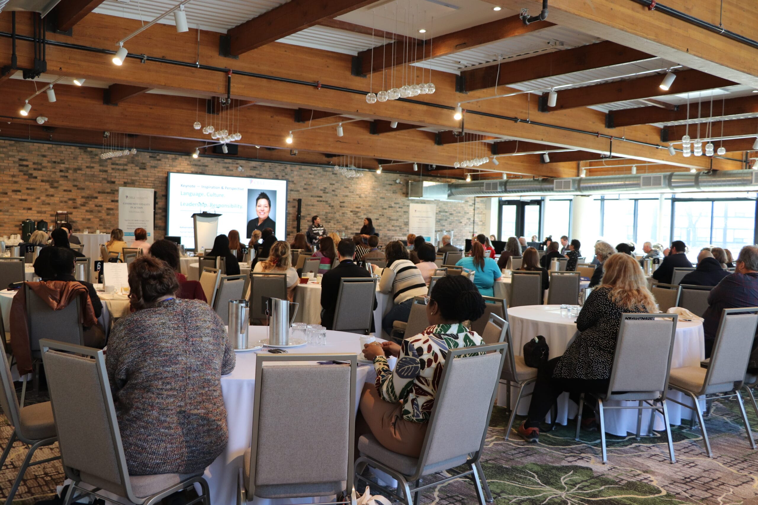 A large conference room with wooden beams filled with seated attendees facing a presenter on stage with a screen display.