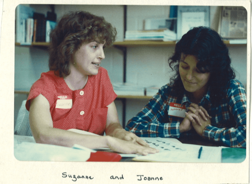 Two women sitting at a table reviewing documents together in a library setting with bookshelves in the background.