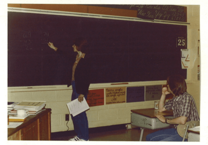 Student writing on classroom blackboard while teacher sits at desk observing, vintage classroom setting.