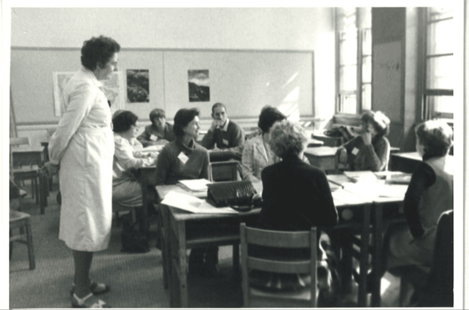 Black and white photograph of a classroom with an instructor in a white coat standing while speaking to seated students around tables.