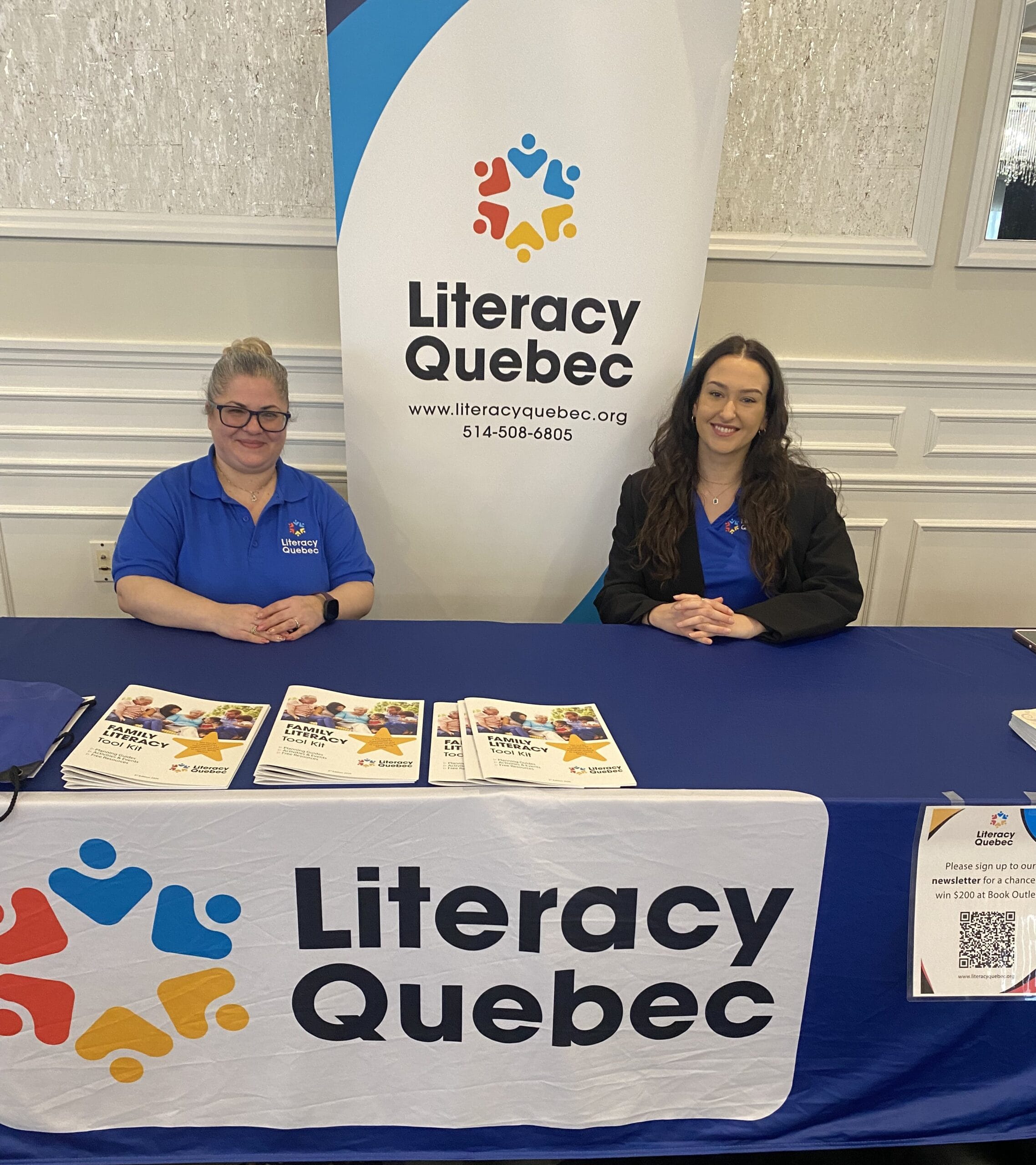 Two women at a Literacy Quebec information booth display promotional materials on a table with branded signage and QR code.