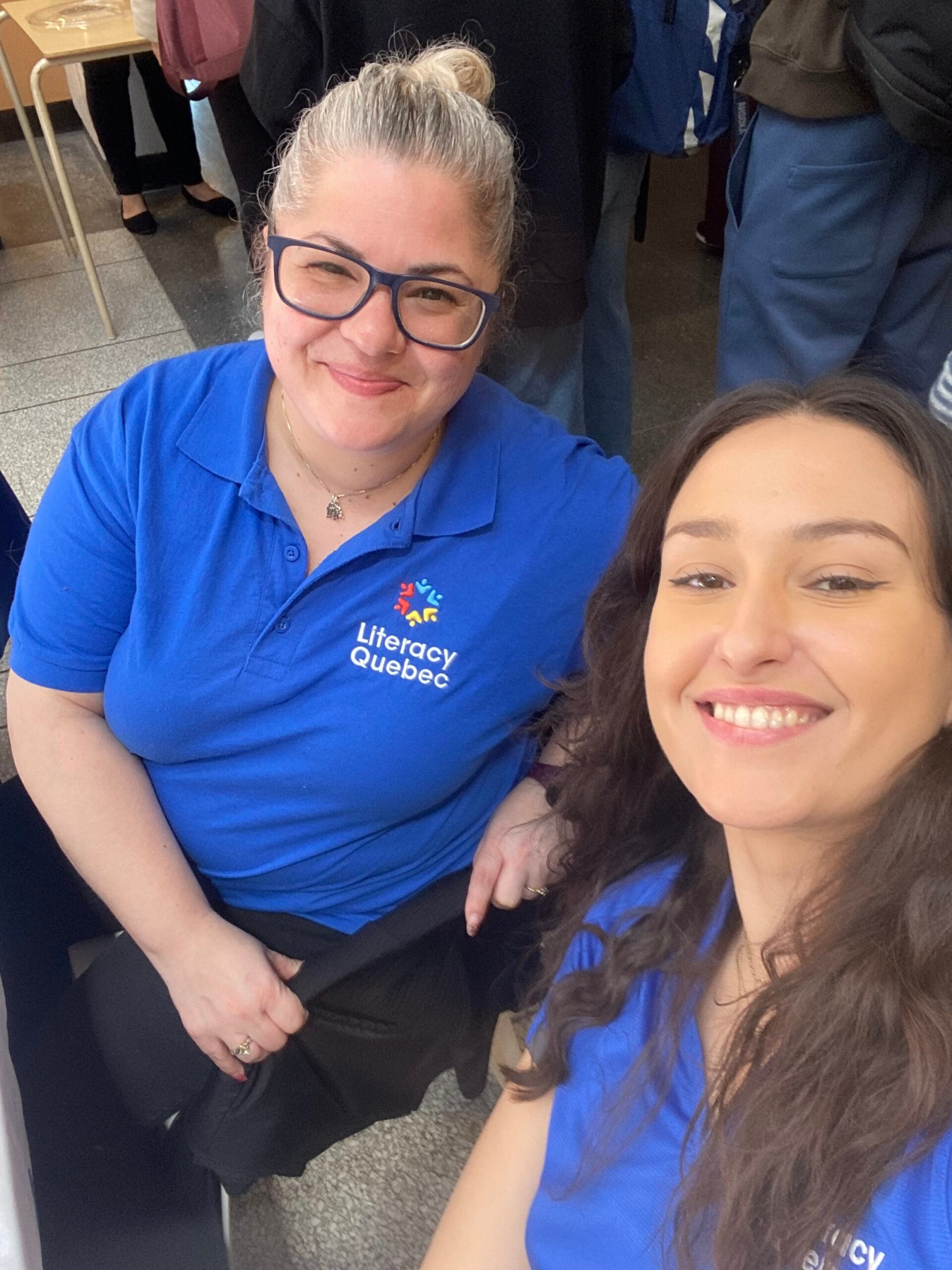 Two women in blue Literacy Quebec polo shirts smiling at a community event indoors.