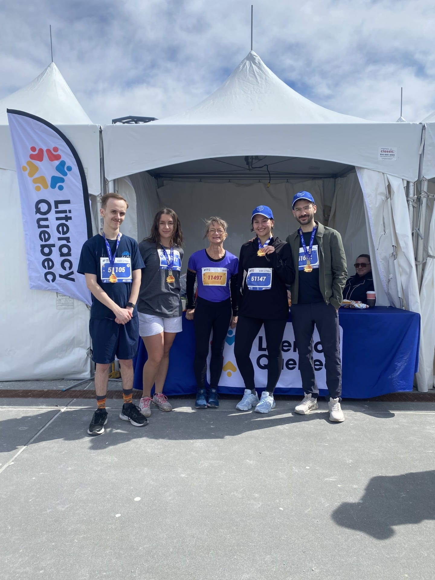 Five participants wearing race bibs stand in front of a white tent at a Literacy Québec event, with a banner and blue table visible.
