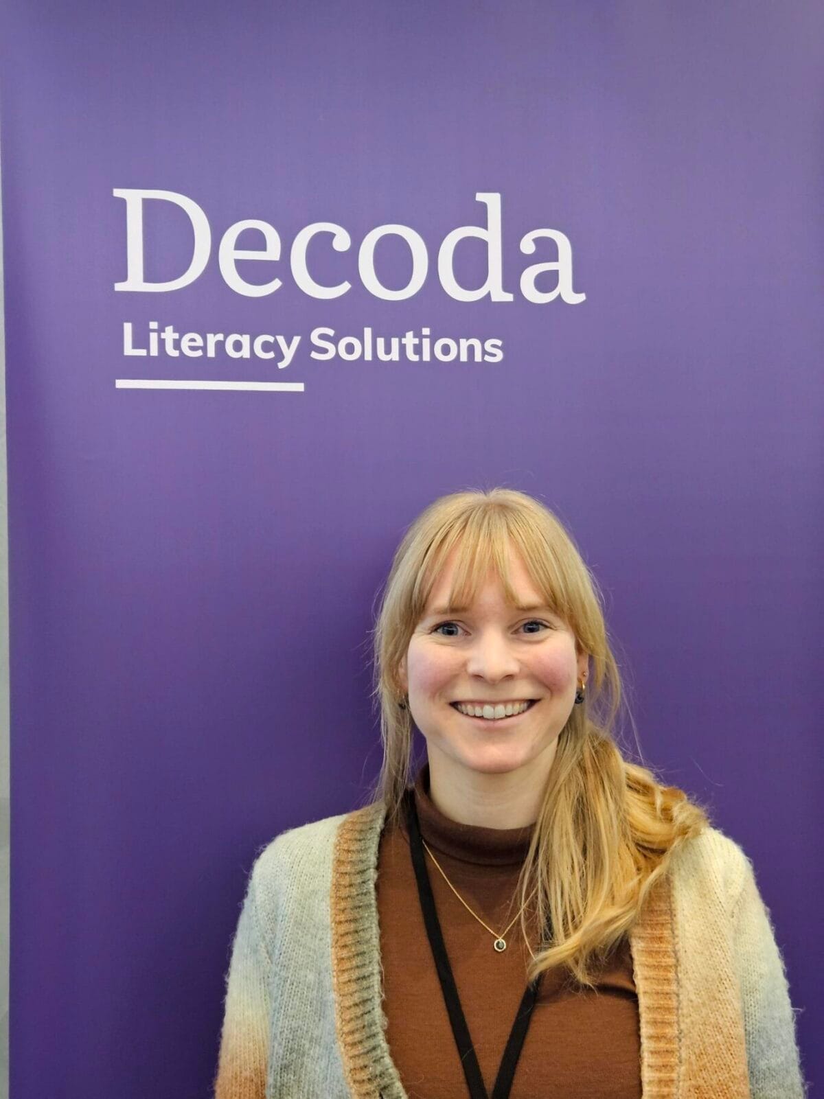 A smiling woman with reddish-blonde hair stands in front of a purple wall displaying "Decoda Literacy Solutions" branding.