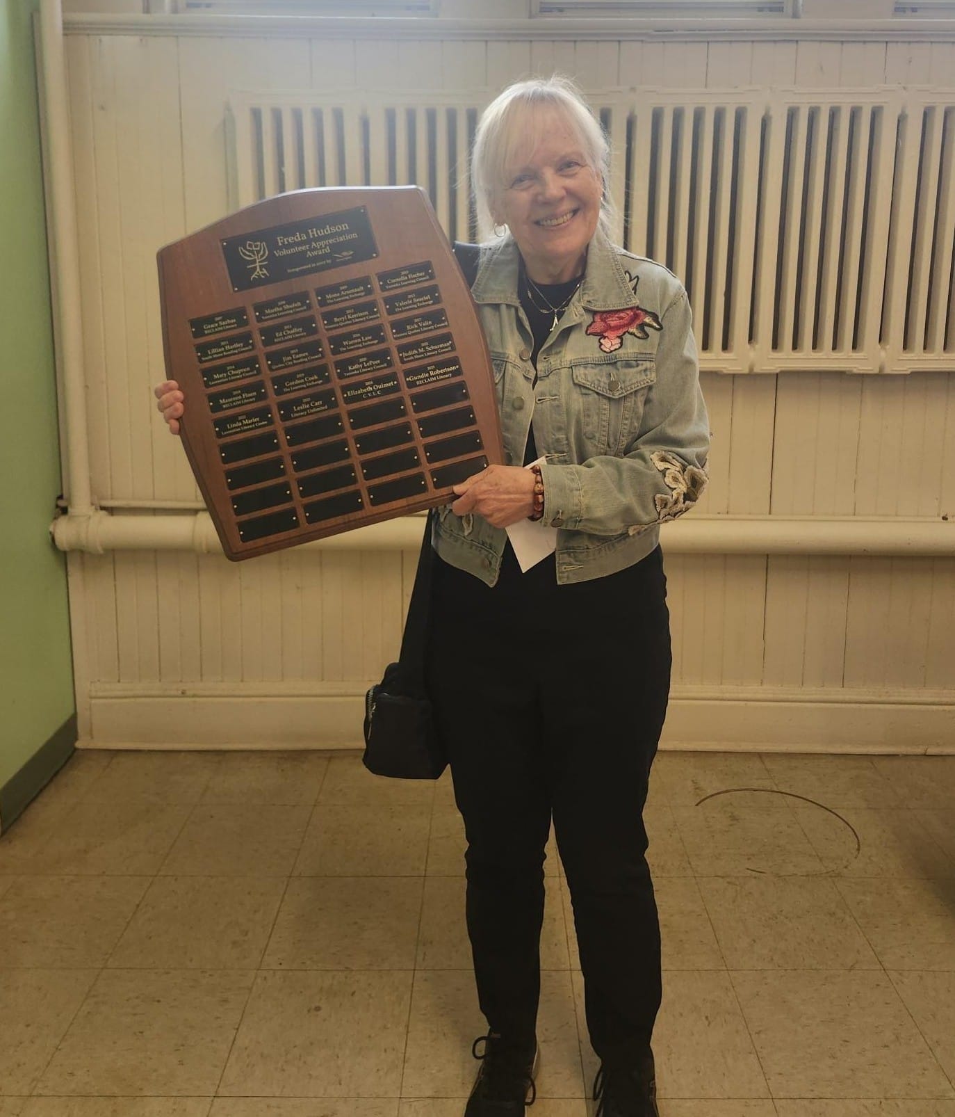 Woman with white hair holding a wooden plaque award with multiple nameplate squares, smiling at camera indoors.